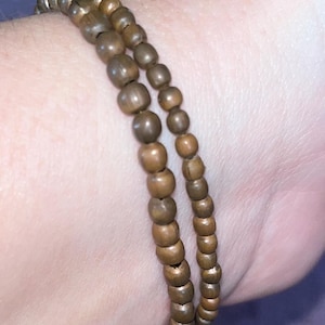 Op de afbeelding: Twee strengen houten kralenarmbanden in verschillende maten. De kralen hebben een warme bruine kleur en lijken aan elkaar geregen te zijn. De armbanden worden om een pols gedragen.