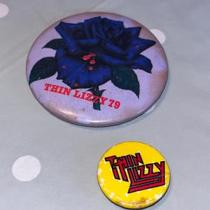 Può includere: Due spille vintage della band Thin Lizzy. Una spilla grande presenta una rosa blu con la scritta "THIN LIZZY 79". L'altra è una spilla gialla più piccola con il logo della band in rosso. Entrambe le spille sono rotonde e mostrano segni di usura.