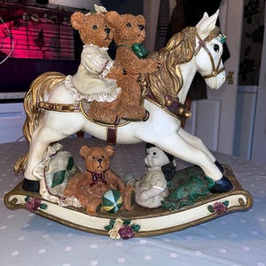 May include: A decorative rocking horse figurine with a white body and gold accents. The horse carries three brown teddy bears. A teddy bear, a doll, and a ball are on the base. The base is decorated with flowers.