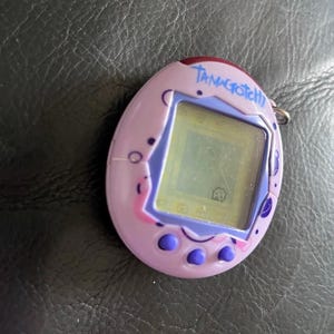 May include: A lavender egg-shaped digital pet device with a purple screen border and three purple buttons at the bottom. The word Tamagotchi is printed in blue script above the screen, which displays a small pixelated character on a light background.