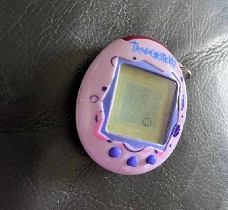 May include: A lavender egg-shaped digital pet device with a purple screen border and three purple buttons at the bottom. The word Tamagotchi is printed in blue script above the screen, which displays a small pixelated character on a light background.