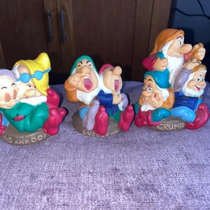 May include: Three ceramic figurines depicting Disney characters. The first shows Dopey and Doc with the text "DOC AND DOPEY". The second shows Sneezy and Happy with the text "SNEEZY AND HAPPY". The third shows Grumpy, Bashful, and Sleepy with the text "GRUMPY".