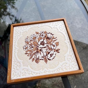 Può includere: Piastrella decorativa quadrata con un motivo floreale marrone e bianco, incorniciata in legno. La piastrella presenta un bouquet centrale di fiori e foglie, con sfondo color crema e bordo decorativo.