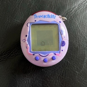 May include: A light purple Tamagotchi digital pet device with a screen displaying a small pixelated character. The egg-shaped plastic casing features dark purple circular patterns, three round buttons below the screen, and the word Tamagotchi.