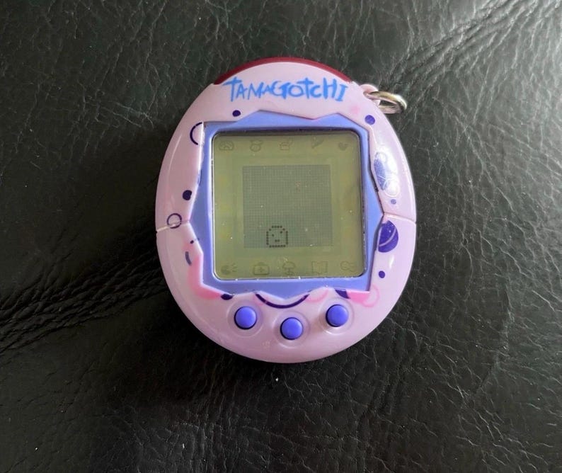 May include: A light purple Tamagotchi digital pet device with a screen displaying a small pixelated character. The egg-shaped plastic casing features dark purple circular patterns, three round buttons below the screen, and the word Tamagotchi.