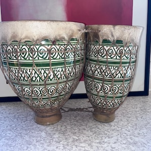 May include: A pair of connected ceramic drums featuring a white and green geometric pattern. The drums have natural skin heads secured with a brown woven rope netting that wraps around the exterior of the glazed ceramic bodies.