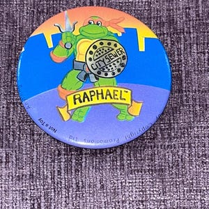 May include: A round pinback button featuring a green cartoon turtle character holding a sai and a circular manhole cover labelled CITY SEWER. The character wears an orange mask and belt. A yellow banner below reads RAPHAEL. Text along the edge reads Not a Toy.