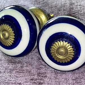May include: Two decorative cabinet knobs. Each knob is a sphere with a navy blue and white striped pattern. The centre of each knob features a gold-coloured, sunburst-shaped design. The knobs have brass-coloured hardware.