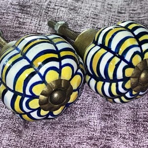 May include: Two decorative ceramic cabinet knobs. Each knob is a rounded shape with a yellow, white, and blue striped pattern. The knobs have a brass-colored base and a threaded metal rod for installation.