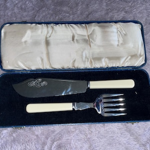 May include: A vintage butter knife and fork set with ivory-colored handles, presented in a blue case with a satin lining. The knife blade has an ornate engraved design. The set is displayed on a dark blue velvet surface.