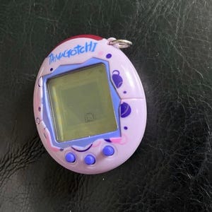 May include: A light purple Tamagotchi digital pet device with a screen displaying a small character. The oval-shaped plastic shell features dark purple and pink circular patterns, three round purple buttons, and a silver metal keychain ring.