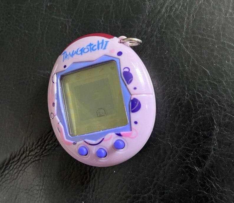 May include: A light purple Tamagotchi digital pet device with a screen displaying a small character. The oval-shaped plastic shell features dark purple and pink circular patterns, three round purple buttons, and a silver metal keychain ring.