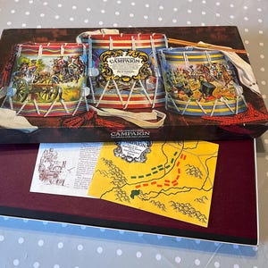 Könnte beinhalten: Eine Vintage-Brettspielbox mit Illustrationen von Trommeln und Kampfszenen. Die Box ist geöffnet und zeigt eine Karte und andere Spielkomponenten. Das Spiel trägt den Titel "Campaign" und hat ein weinrotes Innenleben.