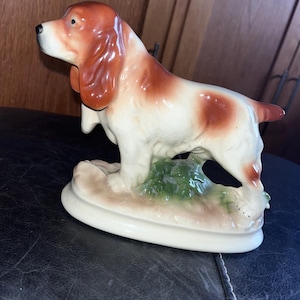 May include: A ceramic dog figurine, featuring a spaniel breed, in a standing pose. The dog has a white body with brown markings on its head, ears, and back. It stands on a cream-colored base with green and brown accents.
