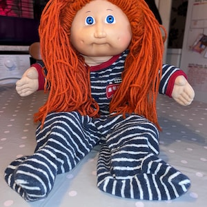 May include: A vintage doll with a vinyl face, blue eyes, and orange yarn hair. The doll is wearing a blue and white striped pajama set with a red trim and a small fire engine graphic on the chest. The doll is sitting on a dotted surface.