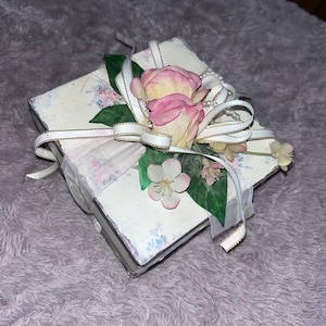 May include: A square decorative box with a floral design, decorated with pink and yellow roses, white ribbons, and faux pearls. The box has a light background with a delicate floral pattern and is tied with a white ribbon with gold trim.
