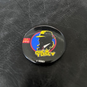 Op de afbeelding: Een ronde, zwarte Dick Tracy-pin met een silhouet van de detective met een gele hoed en een rode en blauwe achtergrond. Het McDonald's-logo is zichtbaar aan de linkerkant. De tekst "Dick Tracy" is geel.