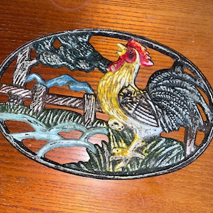 May include: Oval cast iron trivet depicting a rooster crowing in a rural scene. The rooster is yellow, white, and black with a red comb. The background includes a fence, trees, and a stream, all in muted colours.