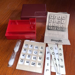 Pode incluir: Um conjunto de jogos de palavras Pocket Boggle, com uma caixa castanha, uma bandeja vermelha, uma grelha de letras bege e uma ampulheta. O jogo inclui peças com letras e instruções para o jogo de palavras de 3 minutos.