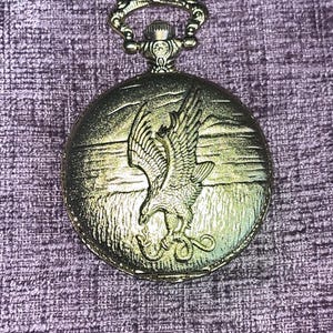 Puede incluir: Un reloj de bolsillo de tono dorado con caja circular que presenta un diseño en relieve de un águila agarrando una serpiente. El reloj tiene un arco decorativo en la parte superior y un acabado metálico texturizado con una escena detallada.