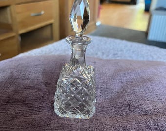 May include: A clear, cut-glass perfume bottle with a crystal stopper. The bottle features a diamond pattern and a tall, pointed stopper. The bottle is on a purple surface.