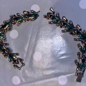 Peut inclure: Un bracelet doré orné d'une série de breloques en forme de grenouilles stylisées avec des détails en émail vert. Le bracelet est présenté non fermé, révélant le mécanisme de fermeture. Les grenouilles sont disposées en motif répétitif.