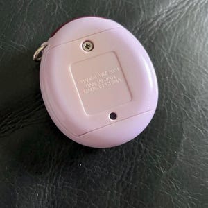 May include: A light purple, oval-shaped electronic handheld game device with a metal keychain loop. The back features a battery compartment cover with the embossed text: &copy;BANDAI WiZ 2001, BANDAI 2004, MADE IN CHINA, and a single visible screw.