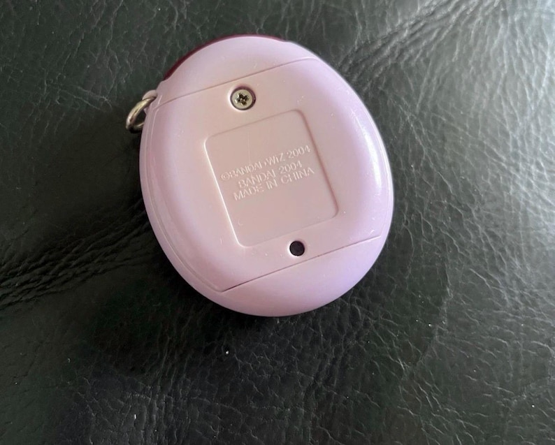 May include: A light purple, oval-shaped electronic handheld game device with a metal keychain loop. The back features a battery compartment cover with the embossed text: &copy;BANDAI WiZ 2001, BANDAI 2004, MADE IN CHINA, and a single visible screw.
