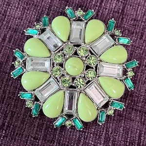 Op de afbeelding: Een ronde broche met een bloemmotief. Het heeft limoengroene, heldere en smaragdgroene edelstenen, gezet in een zilverkleurig metaal. De broche heeft een diameter van ongeveer 5 cm.