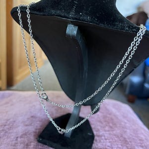 May include: Silver-toned necklace with two strands, displayed on a black velvet bust. The necklace features a delicate chain design with small, round accents. The bust is set on a black stand, against a soft pink surface.