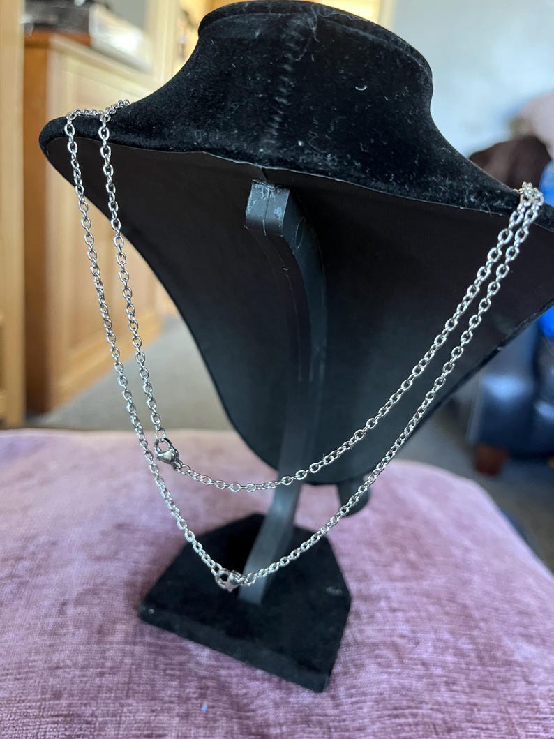 May include: Silver-toned necklace with two strands, displayed on a black velvet bust. The necklace features a delicate chain design with small, round accents. The bust is set on a black stand, against a soft pink surface.