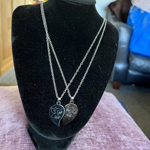 May include: Two silver-toned necklaces displayed on a black mannequin. Each necklace features a pendant shaped like a half-heart with a cartoon character etched on it. The chains are delicate, and the pendants are shiny.