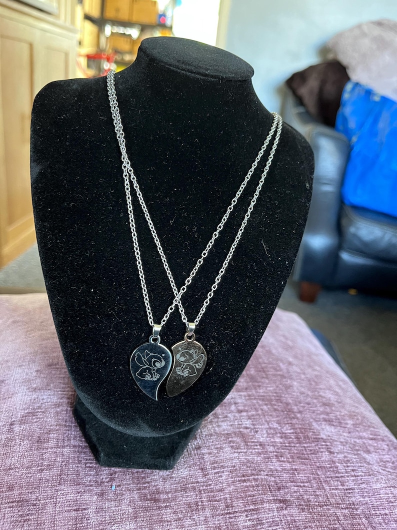 May include: Two silver-toned necklaces displayed on a black mannequin. Each necklace features a pendant shaped like a half-heart with a cartoon character etched on it. The chains are delicate, and the pendants are shiny.