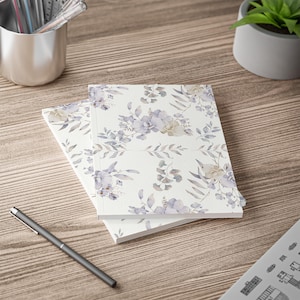 Könnte beinhalten: Zwei Notizbücher mit einem floralen Muster aus Lavendel- und beigefarbenen Blumen und Blättern auf weißem Hintergrund. Die Notizbücher sind auf einer Holzoberfläche gestapelt, mit einem Stift und einem Metallbecher mit Stiften im Hintergrund.