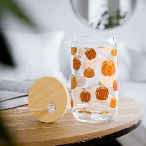 May include: A clear glass can-shaped drinking glass with a bamboo lid and straw. The glass is decorated with orange and peach-coloured pumpkin illustrations. The lid is made of light-coloured bamboo. The glass sits on a wooden table.