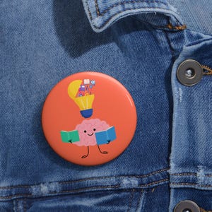 Puede incluir: Un botón redondo y naranja con un cerebro de dibujos animados leyendo libros. Encima del cerebro hay una bombilla con objetos coloridos en su interior. El botón está sobre una chaqueta vaquera azul.