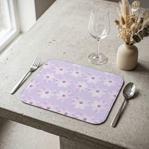 May include: A rectangular lavender placemat with a repeating floral pattern of white and purple flowers. A silver fork and spoon are on either side of the placemat. A wine glass and a small vase with dried flowers are also present.