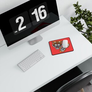 Könnte beinhalten: Ein rotes Mauspad mit einem Cartoon-Tiger-Motiv, eine weiße Maus, eine silberne Tastatur und ein Computermonitor, der die Uhrzeit 14:16 Uhr anzeigt. Die Artikel befinden sich auf einem weißen Schreibtisch.