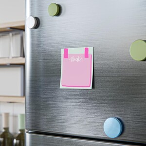 Può includere: Un magnete rosa a forma di lista di cose da fare con la scritta "to do" in corsivo bianco. Il magnete è attaccato a una porta di frigorifero in acciaio inossidabile con accenti rosa a forma di nastro adesivo. Sono visibili anche magneti rotondi verdi e blu.