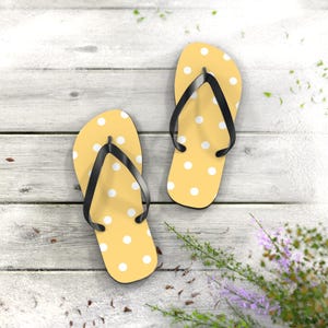 May include: A pair of yellow flip-flops with white polka dots and black straps. The sandals are arranged on a light-colored wooden surface, suggesting a beach or summer theme. The design is simple and cheerful.