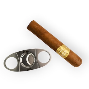 May include: A brown cigar with a gold band next to a silver cigar cutter. The cigar cutter has two holes and a circular blade. The text "Ivan Fox" is engraved on the cutter.