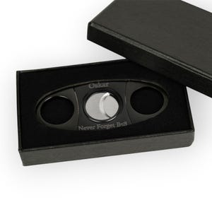 May include: A black cigar cutter with a silver blade, presented in a black box. The cutter has the name "Oskar" engraved on it, along with the phrase "Never Forget Be8". The box is open, revealing the cutter nestled in black velvet.