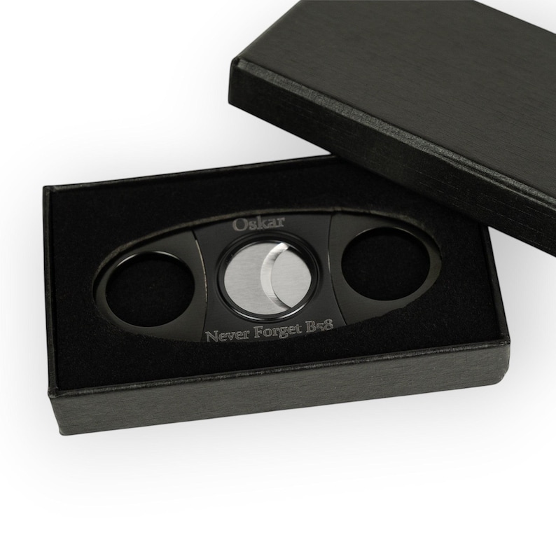 May include: A black cigar cutter with a silver blade, presented in a black box. The cutter has the name "Oskar" engraved on it, along with the phrase "Never Forget Be8". The box is open, revealing the cutter nestled in black velvet.