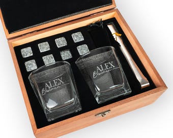 May include: A wooden gift box with a black velvet interior holds two square whiskey glasses engraved with "ALEX Bestman", eight granite whiskey stones, and a pair of stainless steel tongs. The box is made of light brown wood.