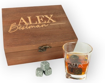 Puede incluir: Una caja de madera con el texto grabado "ALEX Bestman" y un vaso de whisky con cubitos de hielo. Tres piedras de whisky de granito gris están al lado del vaso. La caja tiene un cierre de metal.