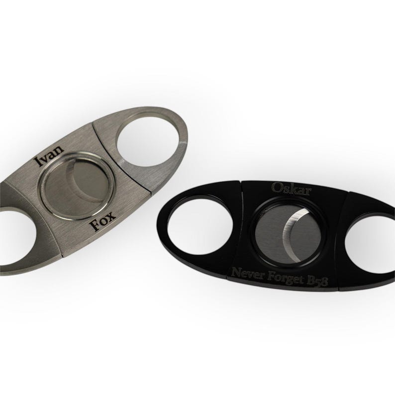 May include: Two cigar cutters with dual blades and finger holes. One is brushed silver engraved with "Ivan" and "Fox". The other is matte black engraved with "Oskar" and "Never Forget B48". Both feature a central circular opening for cutting.