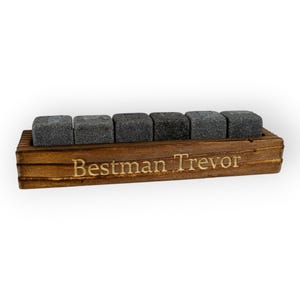 May include: A wooden whiskey stone set with six grey square stones. The stones rest in a stained wooden holder with the engraved text "Bestman Trevor". The set is designed for chilling drinks.