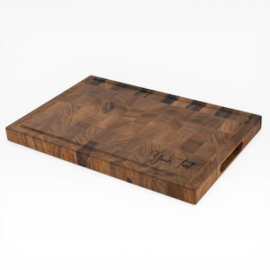 May include: A rectangular end-grain butcher block cutting board made of dark brown wood. It features a juice groove around the edge and a handle cutout. The words "Your Text" are inscribed on the board.