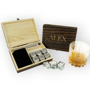 May include: A wooden box set containing stainless steel whiskey stones, a velvet pouch, and tongs. The box is open, revealing the contents, with a second closed box and a glass of amber liquid. The stones are engraved with "AL".