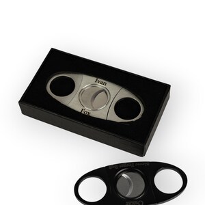 May include: A silver-toned metal cigar cutter engraved with the names Ivan and Fox, presented in a black box, alongside a separate black cigar cutter engraved with the text Never Forget Bc8 and the name Oskar.